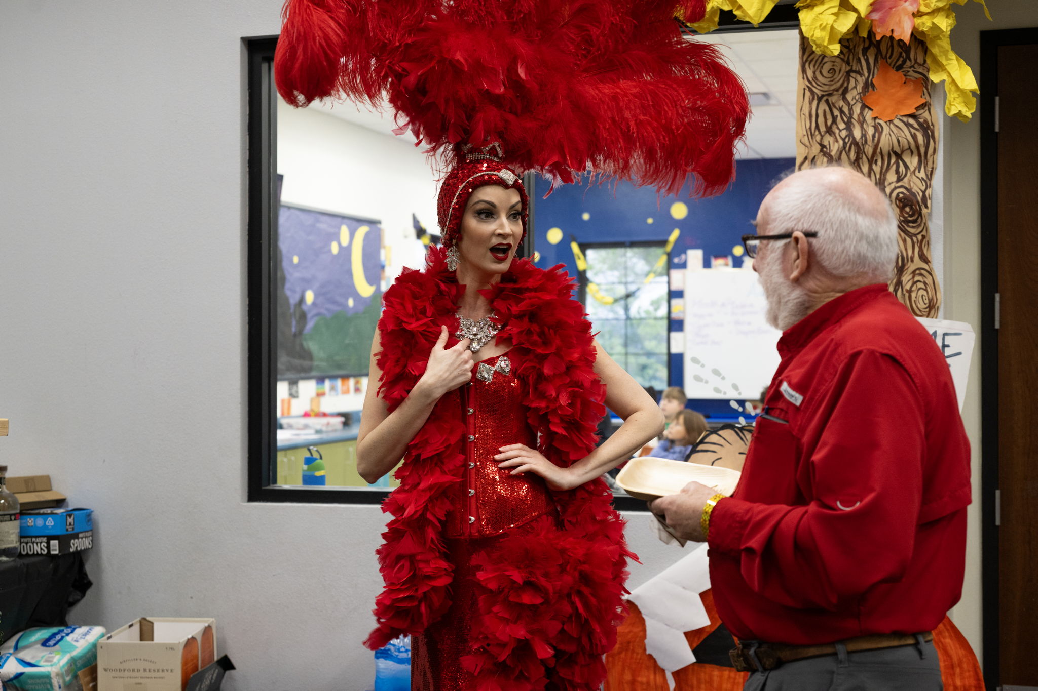 A performer in a vibrant red feathered costume engages in conversation with a man in red, set against a colorful classroom backdrop.