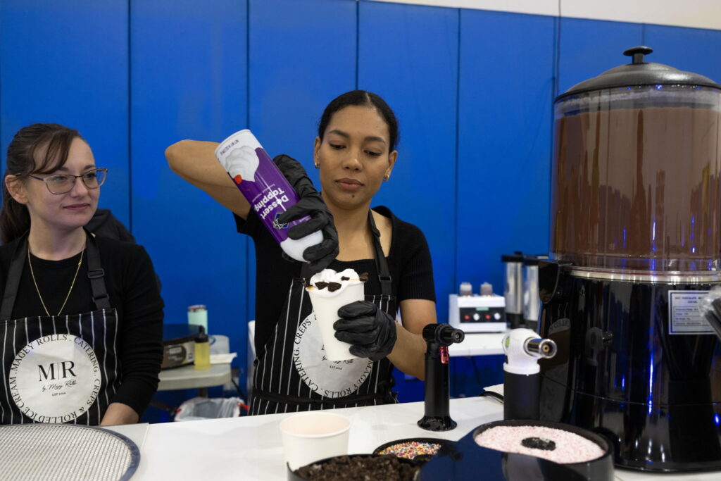 A barista garnishes a dessert cup with whipped cream, while a colleague observes. Chocolate fountain and toppings are visible on the counter.