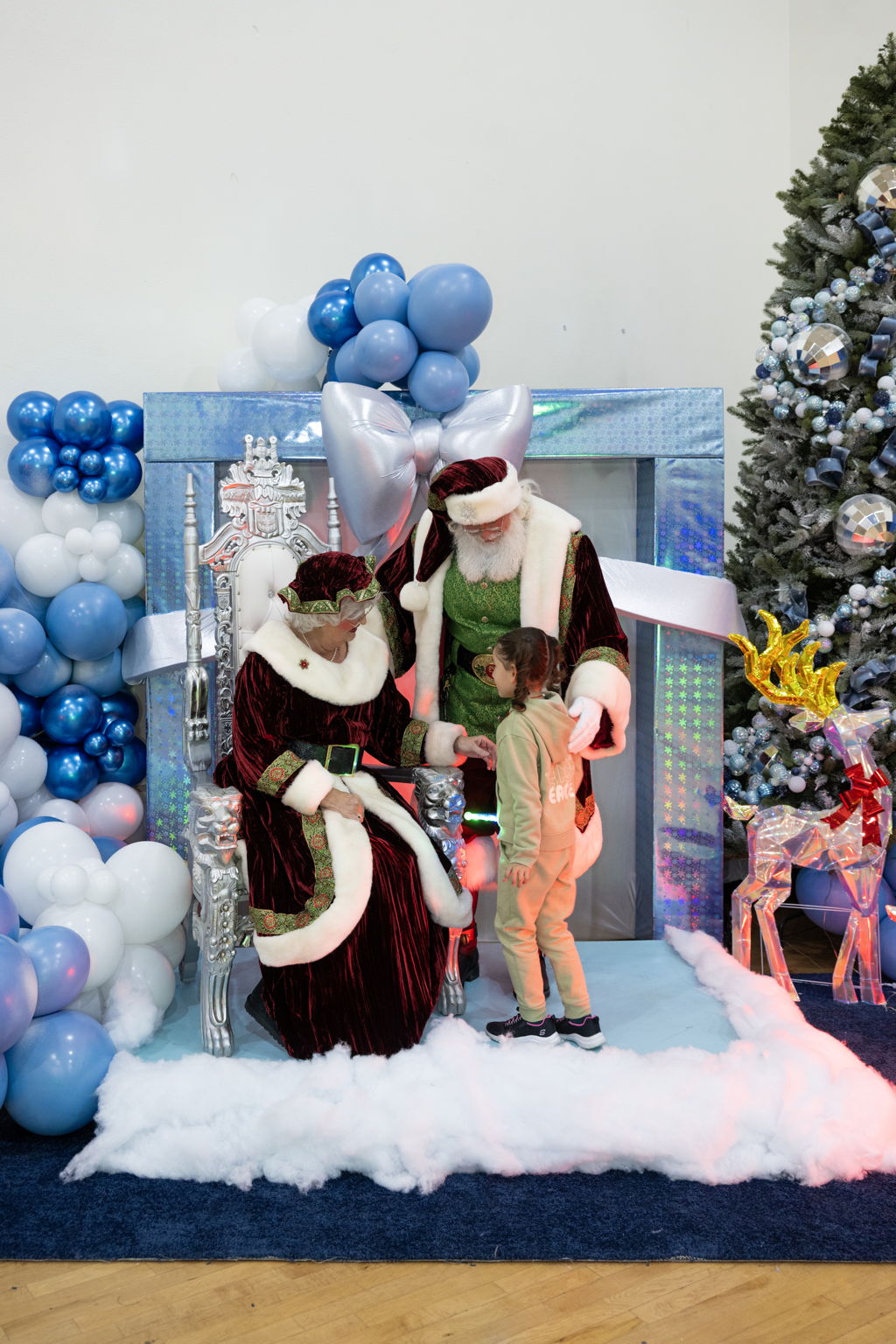 A young girl interacts with Santa and Mrs. Claus, seated against a festive backdrop of blue and white balloons, Christmas decorations, and a tree.