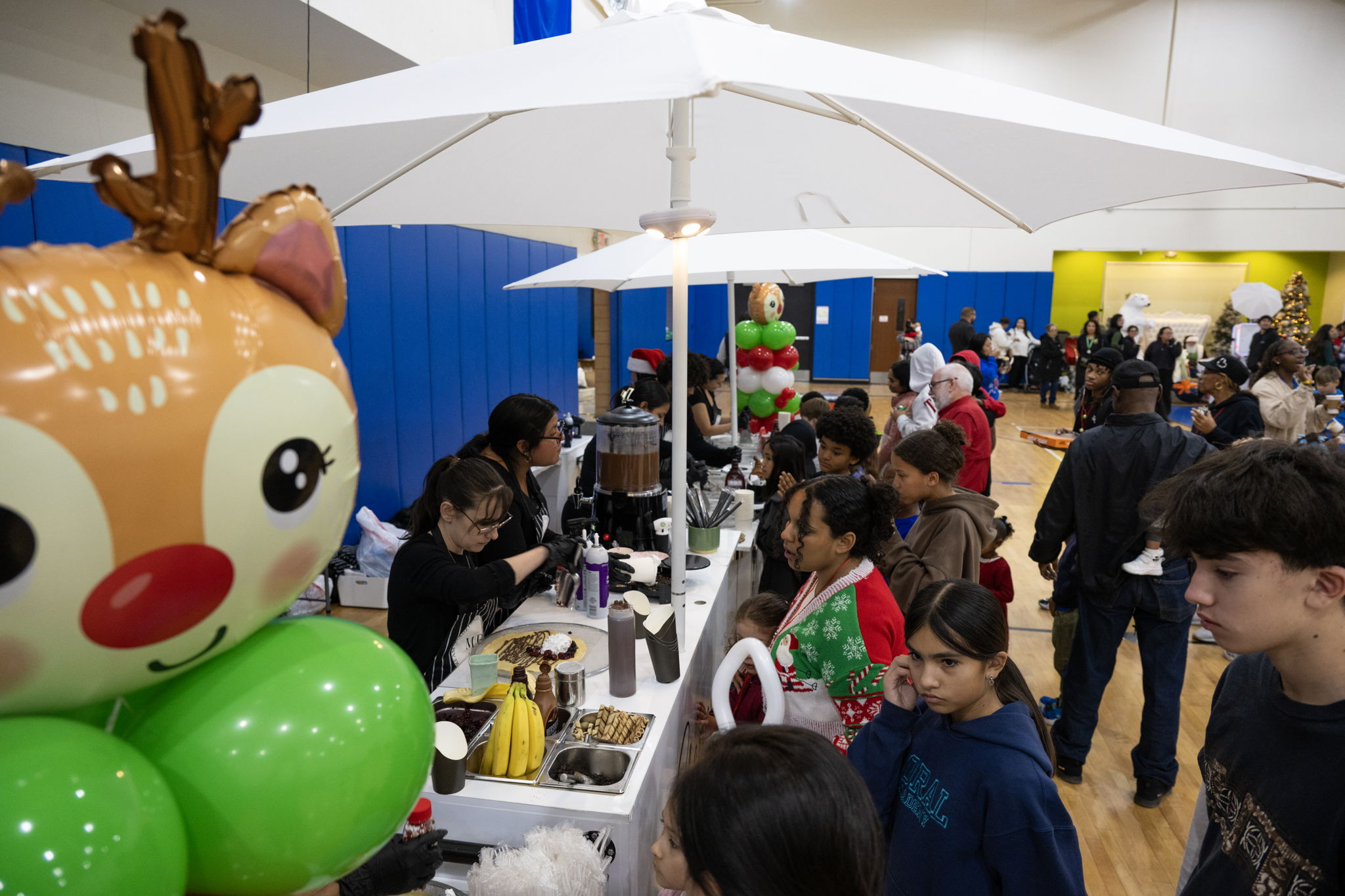 Children and families gather around a festive booth serving treats at a holiday event, featuring a reindeer balloon and decorated treats.